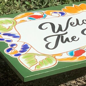 May include: A rectangular green sign featuring a white central panel with the black text "Welcome to the V". The border is decorated with a colorful pattern of orange, blue, and light green stylized floral and leaf motifs on a green background.