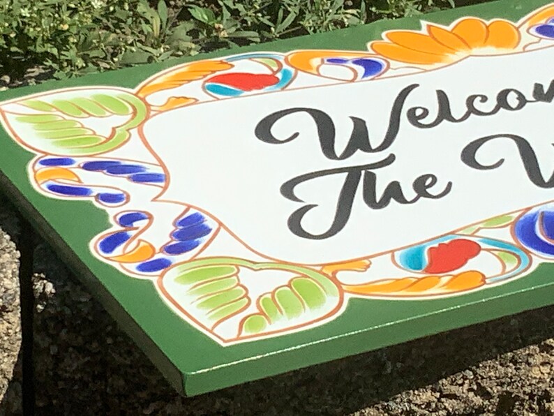 May include: A rectangular green sign featuring a white central panel with the black text "Welcome to the V". The border is decorated with a colorful pattern of orange, blue, and light green stylized floral and leaf motifs on a green background.