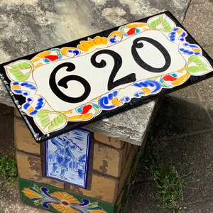 May include: A rectangular ceramic house number plaque featuring the black digits 620 centered on a cream background. The border is decorated with a colorful hand-painted floral and scroll pattern in shades of green, blue, orange, and yellow.