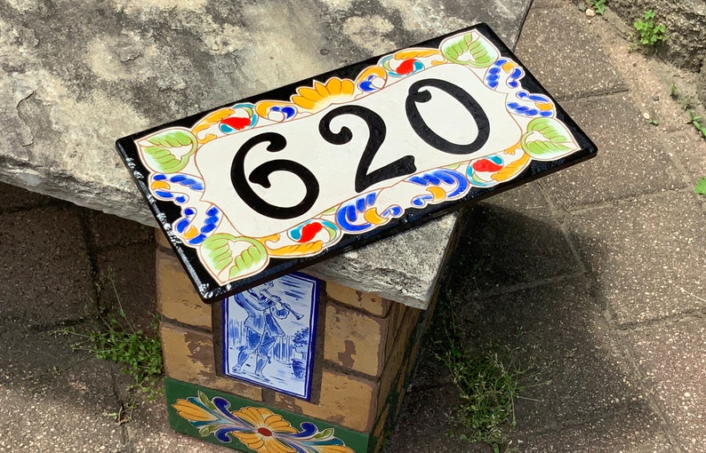 May include: A rectangular ceramic house number plaque featuring the black digits 620 centered on a cream background. The border is decorated with a colorful hand-painted floral and scroll pattern in shades of green, blue, orange, and yellow.