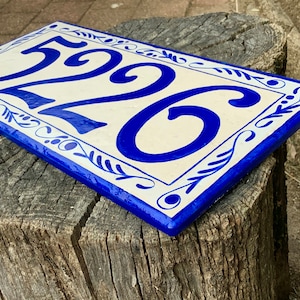 May include: A rectangular ceramic house number plaque featuring the digits 5226 in a bold, glossy blue glaze against a cream-colored background. The plaque is framed by a decorative blue border with hand-painted leaf and vine motifs.
