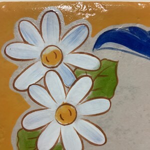 May include: A close-up of a ceramic tile with a painted design of two white daisies with yellow centers and green leaves. The background is a light brown color.