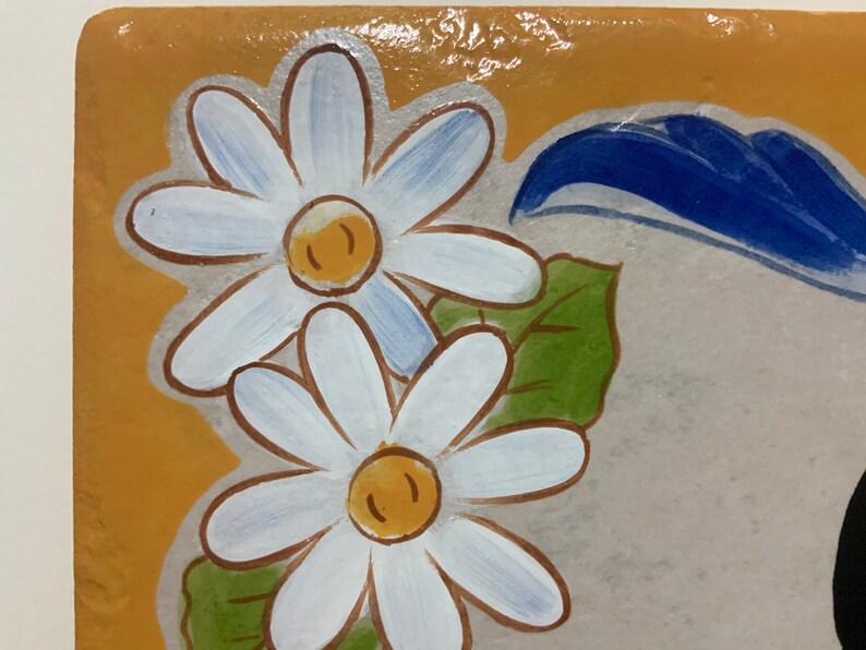 May include: A close-up of a ceramic tile with a painted design of two white daisies with yellow centers and green leaves. The background is a light brown color.