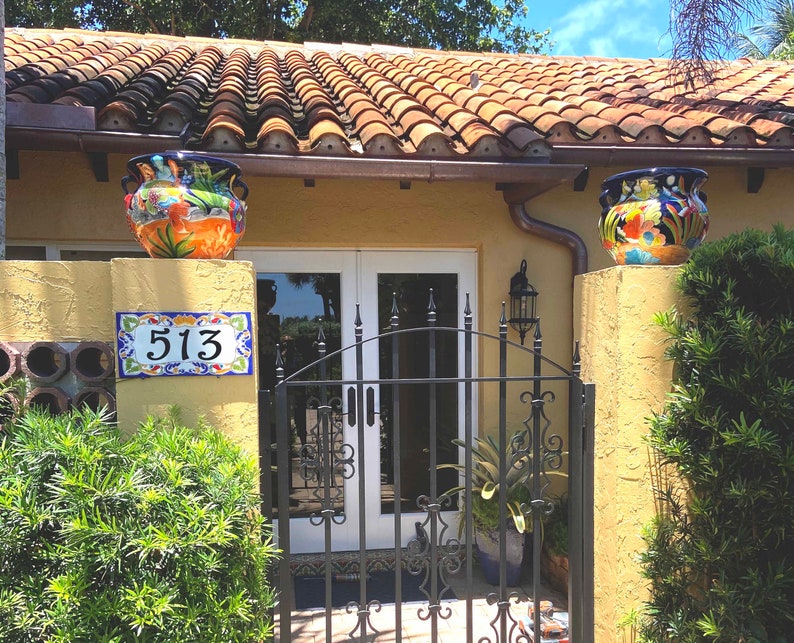 May include: A house entrance featuring a yellow stucco wall with a ceramic tile address plaque displaying the number 513. Two colorful hand-painted ceramic planters with floral motifs sit atop the pillars flanking a black wrought iron gate.