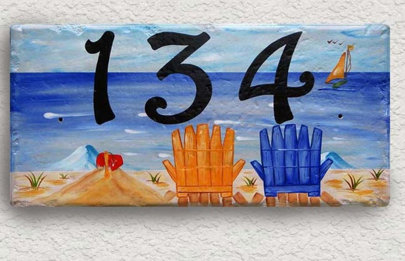 May include: A painted wooden sign with a beach scene and the number 134. The sign features two beach chairs, a sailboat, and a blue ocean.