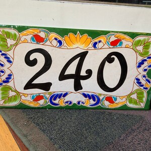 May include: A rectangular ceramic house number plaque featuring the black digits 240 centered on a cream background. The border is decorated with a green frame and colorful painted floral motifs in yellow, blue, and red accents throughout.