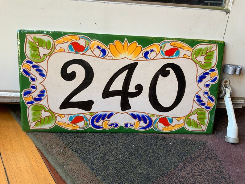 May include: A rectangular ceramic house number plaque featuring the black digits 240 centered on a cream background. The border is decorated with a green frame and colorful painted floral motifs in yellow, blue, and red accents throughout.