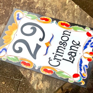 May include: A white ceramic house number sign with black lettering and colorful floral designs. The number 29 is displayed prominently, along with the street name "Crimson Lane".