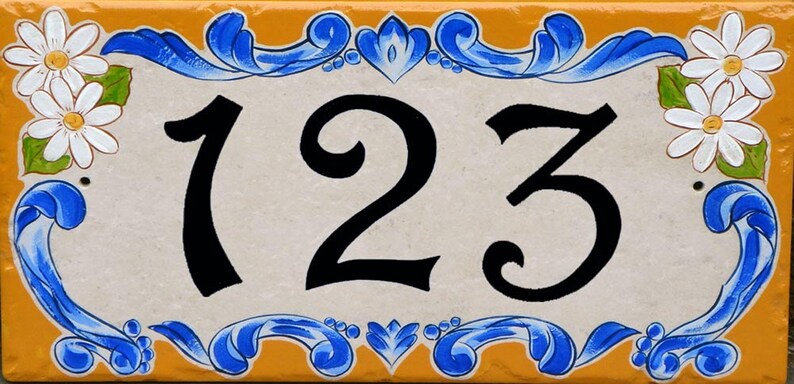 May include: A white ceramic house number sign with blue and yellow floral accents. The number 123 is painted in black.