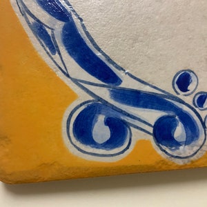 May include: A close-up of a yellow and white ceramic tile with a blue and white design. The design features a swirling pattern with blue accents and white outlines.
