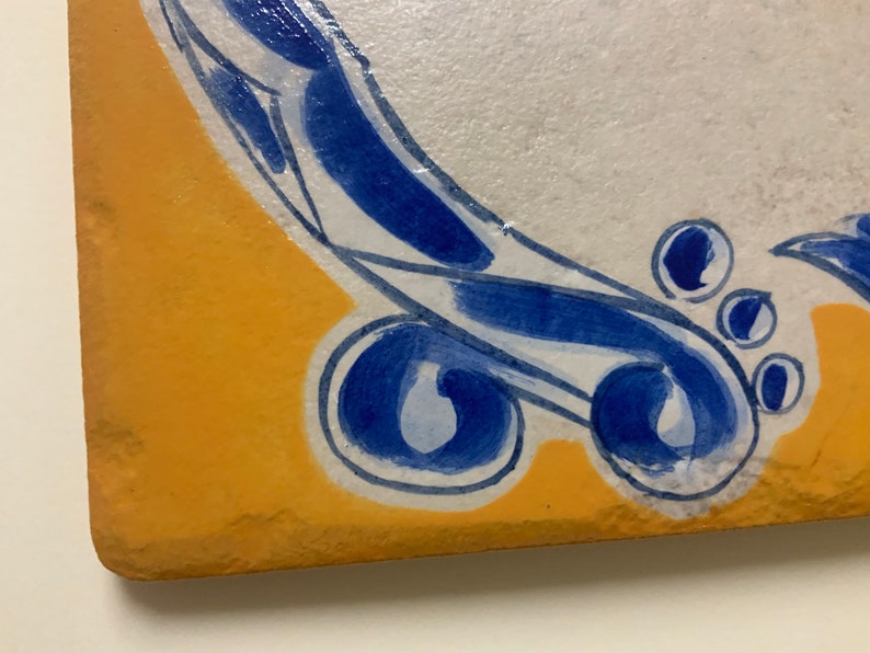 May include: A close-up of a yellow and white ceramic tile with a blue and white design. The design features a swirling pattern with blue accents and white outlines.