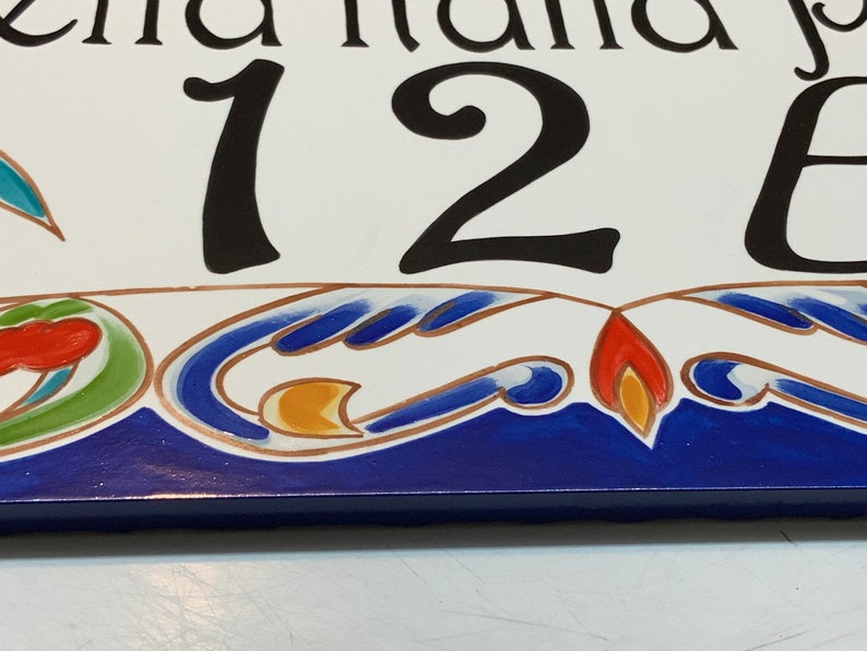 May include: A white ceramic house number sign with the number 126 painted in black. The sign has a blue and white border with a decorative design in red, orange, yellow, and green.
