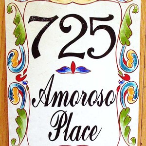 May include: A white ceramic sign with a black painted number 725 and the words "Amoroso Place" in black script. The sign is decorated with colorful floral designs.
