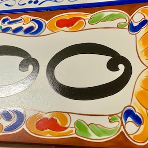 May include: Close-up of a decorative ceramic tile with a white background and a large black letter "O". The tile is framed with a brown border and colorful floral accents in blue, orange, red, and green.