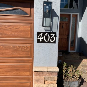 May include: A black metal house number sign with white numbers reading "403" mounted on a light gray stucco wall. A brown wooden garage door is to the left of the sign. A stone pillar is to the right of the sign.
