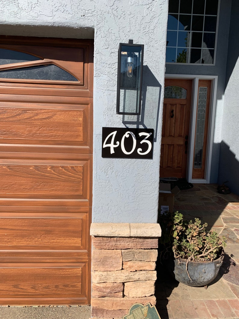 May include: A black metal house number sign with white numbers reading "403" mounted on a light gray stucco wall. A brown wooden garage door is to the left of the sign. A stone pillar is to the right of the sign.