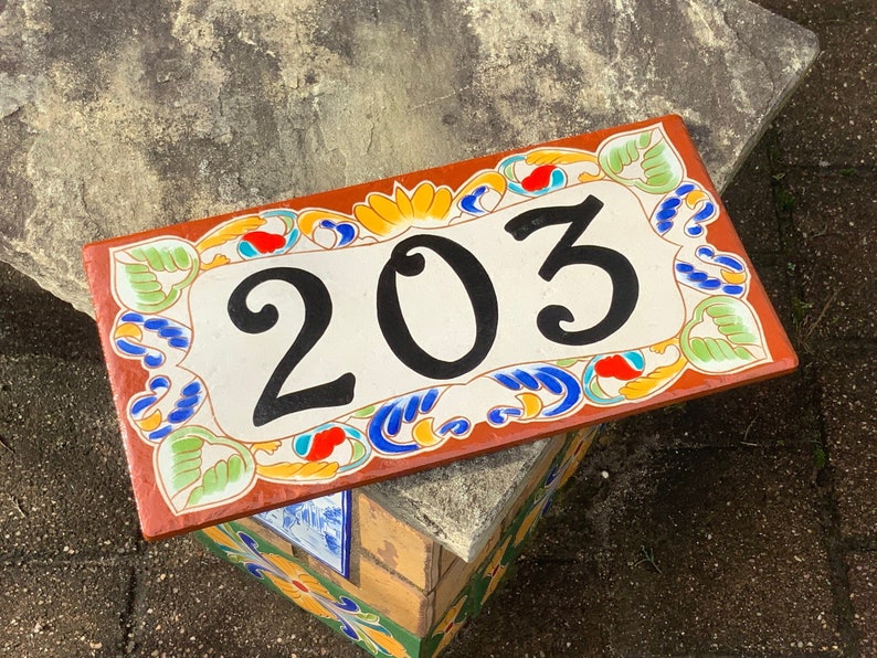 May include: A terracotta-colored ceramic house number sign with a white background and black numbers. The sign reads "203" and is decorated with colorful floral designs.