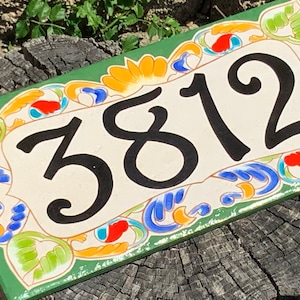 May include: A rectangular ceramic house number plaque featuring the black digits 3812 centered on a cream background. The border is decorated with a hand-painted floral pattern in vibrant shades of green, orange, red, and blue on a green base.