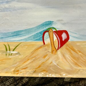 May include: A red bucket and wooden shovel are partially buried in a mound of sand. The background shows a blue wave and a light blue sky.