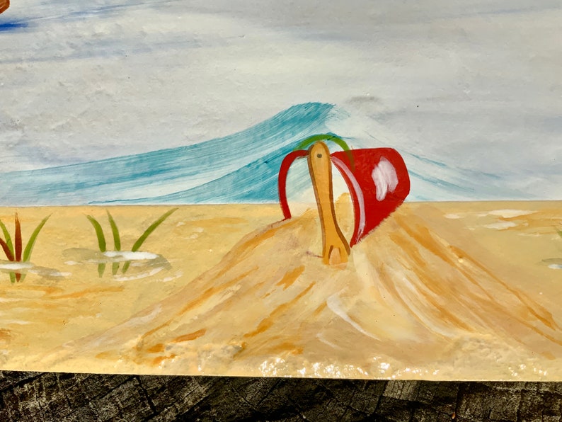 May include: A red bucket and wooden shovel are partially buried in a mound of sand. The background shows a blue wave and a light blue sky.