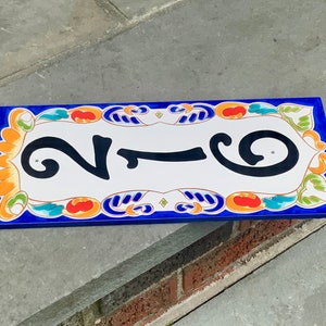 May include: Rectangular ceramic house number plaque with the number 216 in black against a white background. The plaque has a blue border and is decorated with colorful floral and scroll designs in orange, green, and blue.