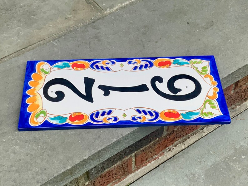 May include: Rectangular ceramic house number plaque with the number 216 in black against a white background. The plaque has a blue border and is decorated with colorful floral and scroll designs in orange, green, and blue.