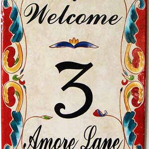 May include: Welcome sign with a red background and colorful floral designs. The sign says "Welcome", "3", and "Amore Lane".