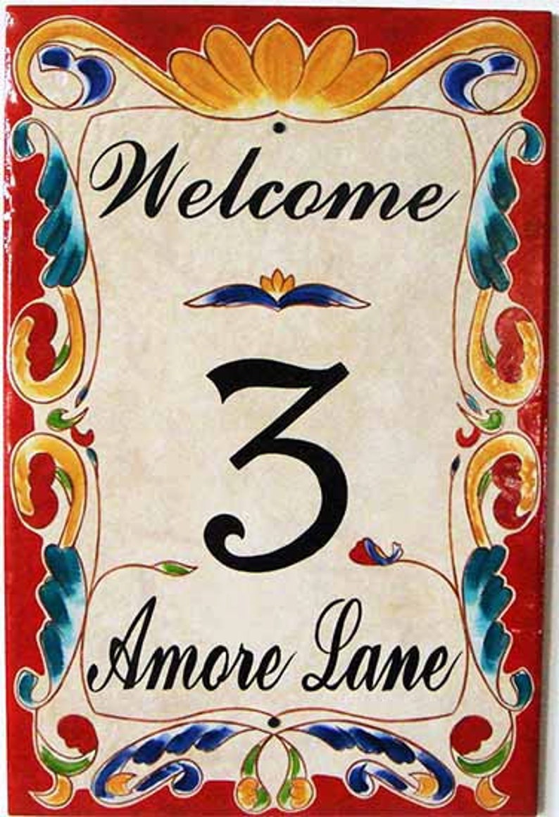May include: Welcome sign with a red background and colorful floral designs. The sign says "Welcome", "3", and "Amore Lane".