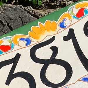May include: A rectangular ceramic house number plaque featuring the black digits 3819 on a cream background. The border is decorated with hand-painted floral and scroll motifs in vibrant shades of orange, blue, red, and green on a white base.