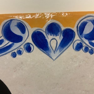 May include: A white ceramic tile with a blue and yellow border. The border features a stylized floral design with blue and white swirls and dots.