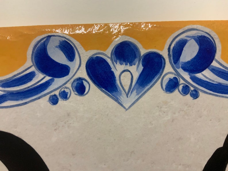 May include: A white ceramic tile with a blue and yellow border. The border features a stylized floral design with blue and white swirls and dots.
