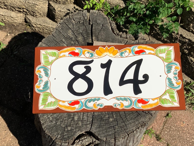 May include: A rectangular house number plaque with a white center featuring the black digits 814. The border is decorated with a colorful floral and leaf pattern in shades of green, orange, and teal against a solid brown background.