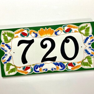 May include: A rectangular house number plaque featuring the black digits 720 centered on a white background. The border is painted in a deep green with decorative floral and leaf motifs in shades of orange, blue, and light green.