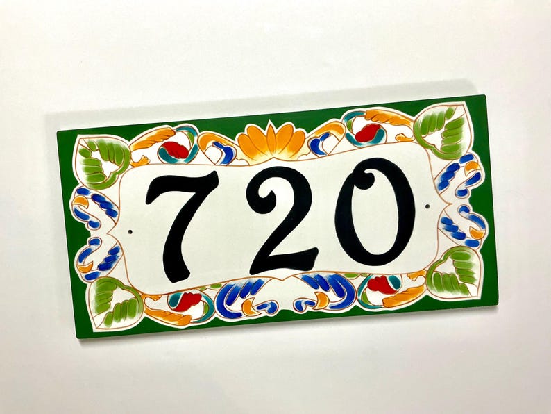 May include: A rectangular house number plaque featuring the black digits 720 centered on a white background. The border is painted in a deep green with decorative floral and leaf motifs in shades of orange, blue, and light green.