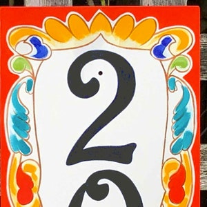 May include: A rectangular, hand-painted house number sign with the number "23" in bold black font. The sign has a white background with a decorative border in orange, yellow, blue, and green. The sign is likely made of ceramic.