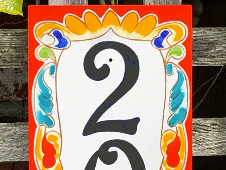 May include: A rectangular, hand-painted house number sign with the number "23" in bold black font. The sign has a white background with a decorative border in orange, yellow, blue, and green. The sign is likely made of ceramic.
