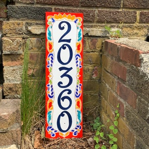 May include: A rectangular address sign with the number "20360" in navy blue against a white background. The sign has a red border with colorful floral designs. The sign is placed against a brick wall.