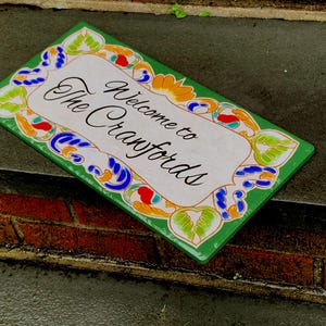 May include: A rectangular ceramic sign with a white center and a green border featuring colorful floral and leaf patterns in blue, orange, and light green. The text "Welcome to The Cranfords" is printed in black script in the center of the sign.