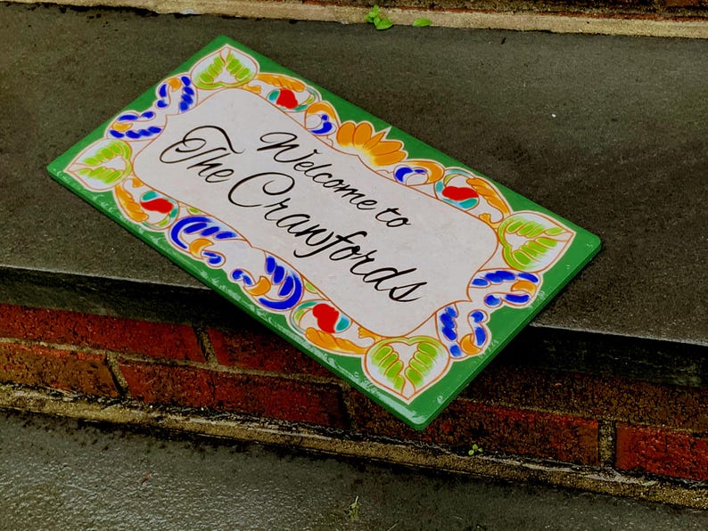 May include: A rectangular ceramic sign with a white center and a green border featuring colorful floral and leaf patterns in blue, orange, and light green. The text "Welcome to The Cranfords" is printed in black script in the center of the sign.