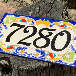 May include: A rectangular ceramic house number plaque featuring the black digits 7280 centered on a cream background. The border is decorated with a vibrant hand-painted pattern of blue, orange, and light green floral and leaf motifs.