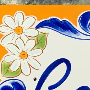 May include: A close-up of a hand-painted ceramic tile with a bright orange background. The tile features two white daisies with yellow centers and green leaves. The tile also has a blue and white floral design in the lower right corner.