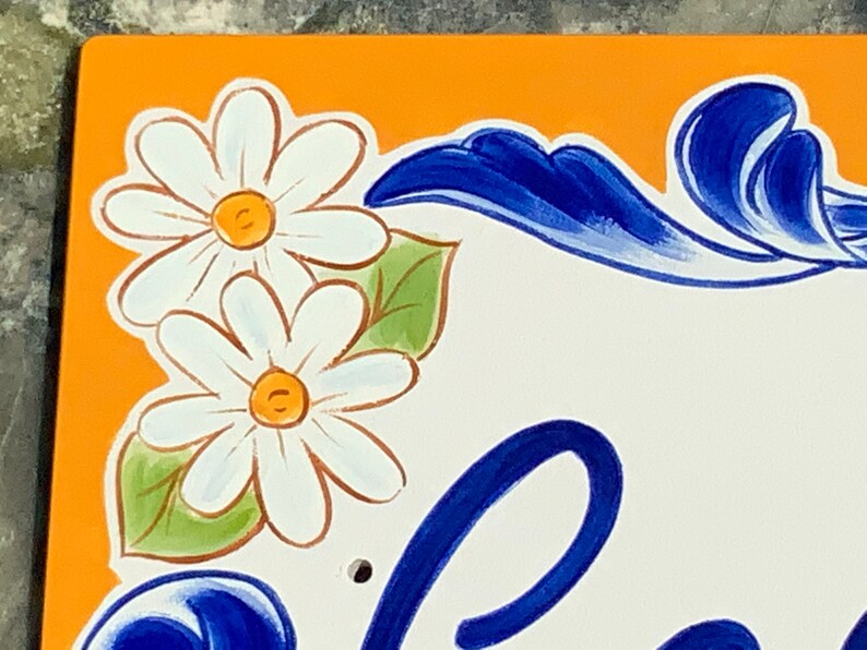 May include: A close-up of a hand-painted ceramic tile with a bright orange background. The tile features two white daisies with yellow centers and green leaves. The tile also has a blue and white floral design in the lower right corner.