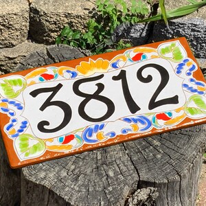 May include: A rectangular ceramic house number plaque featuring the black digits 3812 centered on a white background. The border is decorated with a hand-painted floral pattern in vibrant shades of orange, yellow, blue, and light green.