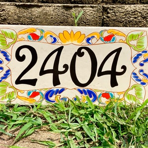 May include: A rectangular ceramic house number plaque featuring the black digits 2404 centered on a cream background. The border is decorated with a colorful hand-painted pattern of green leaves, blue swirls, and orange accents on all four sides.