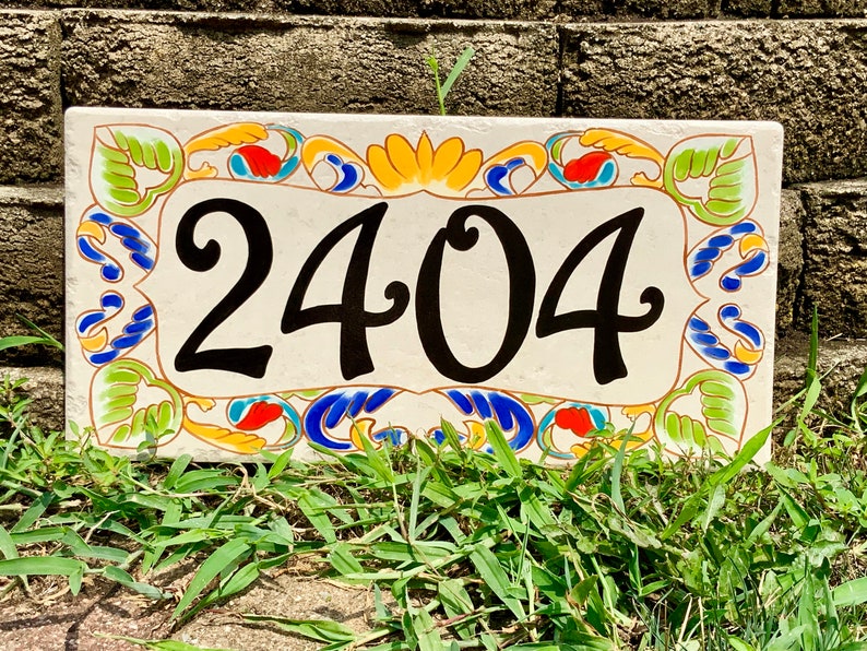 May include: A rectangular ceramic house number plaque featuring the black digits 2404 centered on a cream background. The border is decorated with a colorful hand-painted pattern of green leaves, blue swirls, and orange accents on all four sides.