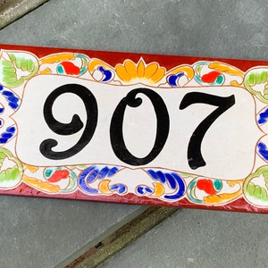 May include: A rectangular ceramic house number plaque featuring the black digits 907 centered on a cream-colored background. The border is decorated with a hand-painted pattern of green leaves, orange floral accents, and blue swirls on a dark red base.