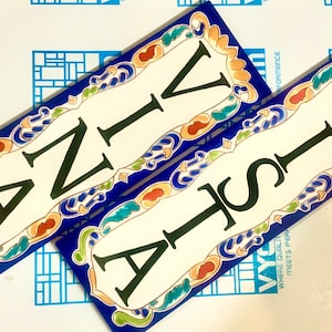 May include: Two rectangular ceramic tiles with the letters "ANIV" and "VISTA" in black. The tiles have a white background with a blue border and decorative floral accents in orange, green, and yellow.