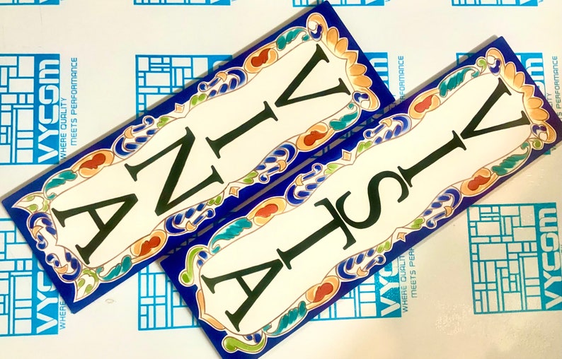 May include: Two rectangular ceramic tiles with the letters "ANIV" and "VISTA" in black. The tiles have a white background with a blue border and decorative floral accents in orange, green, and yellow.