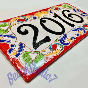 May include: A rectangular ceramic house number plaque featuring the black painted digits 2016 centered on a white background. The border is decorated with hand-painted floral and scroll patterns in vibrant red, blue, green, and yellow colors.