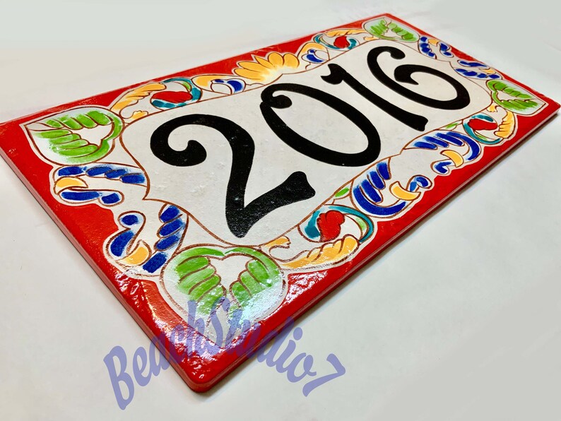 May include: A rectangular ceramic house number plaque featuring the black painted digits 2016 centered on a white background. The border is decorated with hand-painted floral and scroll patterns in vibrant red, blue, green, and yellow colors.